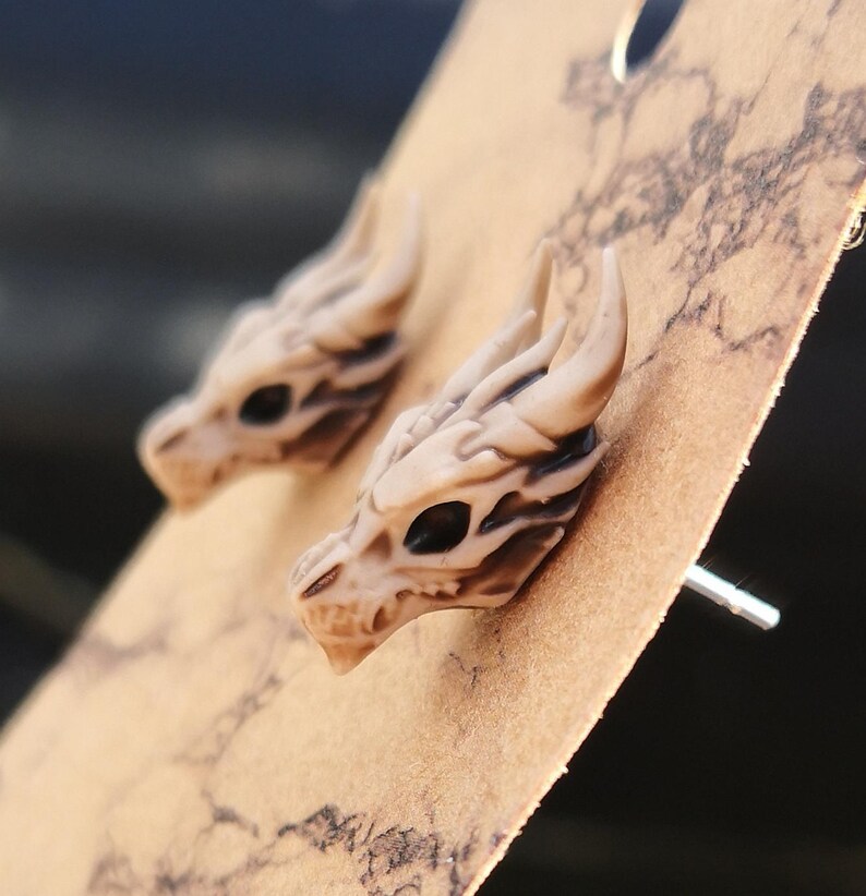 May include: A pair of beige dragon skull stud earrings. The earrings are made of a smooth, textured material and have a detailed design.