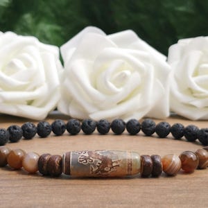 May include: A brown and black lava rock bracelet with a brown agate bead in the centre. The bracelet is on a wooden surface.