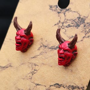 May include: A pair of red devil head stud earrings with brown horns. The earrings are on a tan card with a black hole at the top. The devil heads have an open mouth and detailed facial features.