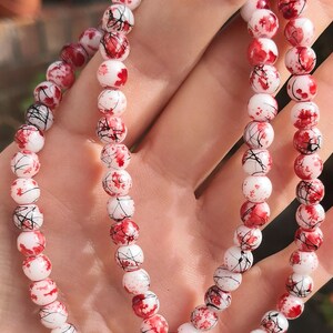 May include: A beaded bracelet featuring round white beads with a decorative pattern of red paint splatters and thin black lines. The beads are strung together in a continuous loop, creating a circular design with a high-gloss finish throughout.
