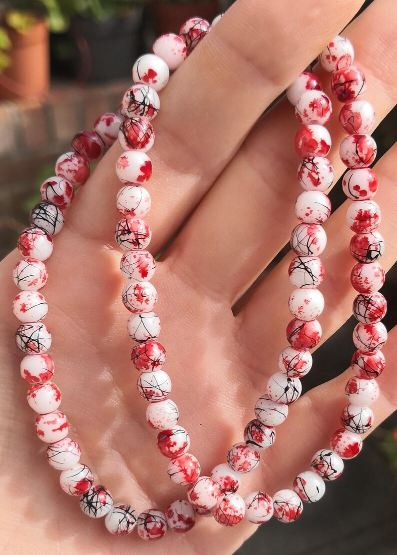 May include: A beaded bracelet featuring round white beads with a decorative pattern of red paint splatters and thin black lines. The beads are strung together in a continuous loop, creating a circular design with a high-gloss finish throughout.