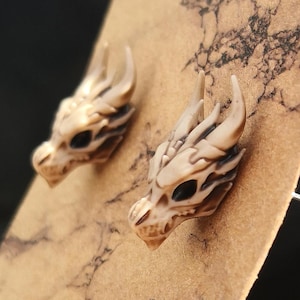 May include: A pair of dragon skull earrings made of white resin. The earrings are displayed on a brown background.