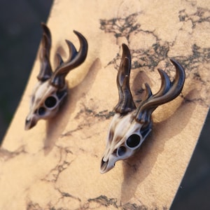 May include: A pair of deer skull earrings with brown antlers. The earrings are made of a white and brown material and have black eyes.