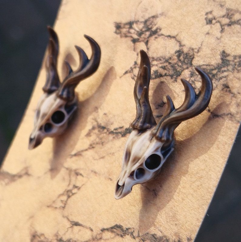 May include: A pair of deer skull earrings with brown antlers. The earrings are made of a white and brown material and have black eyes.