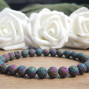 May include: A bracelet made of iridescent lava rock beads, displaying a range of colours including purple, blue, and green. The bracelet is presented in front of white roses.