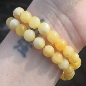 May include: Two bracelets with light yellow, round beads. The bracelets are worn on a wrist, with a tattoo visible on the skin. The beads have a smooth, polished appearance, and the colour varies slightly.