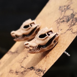 May include: A pair of small, beige alligator skull earrings. The earrings are made of polymer clay and have a realistic look. They are perfect for anyone who loves unique and edgy jewellery.