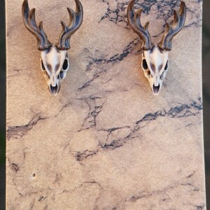 May include: A pair of brown and white deer skull earrings. The earrings are on a brown background.
