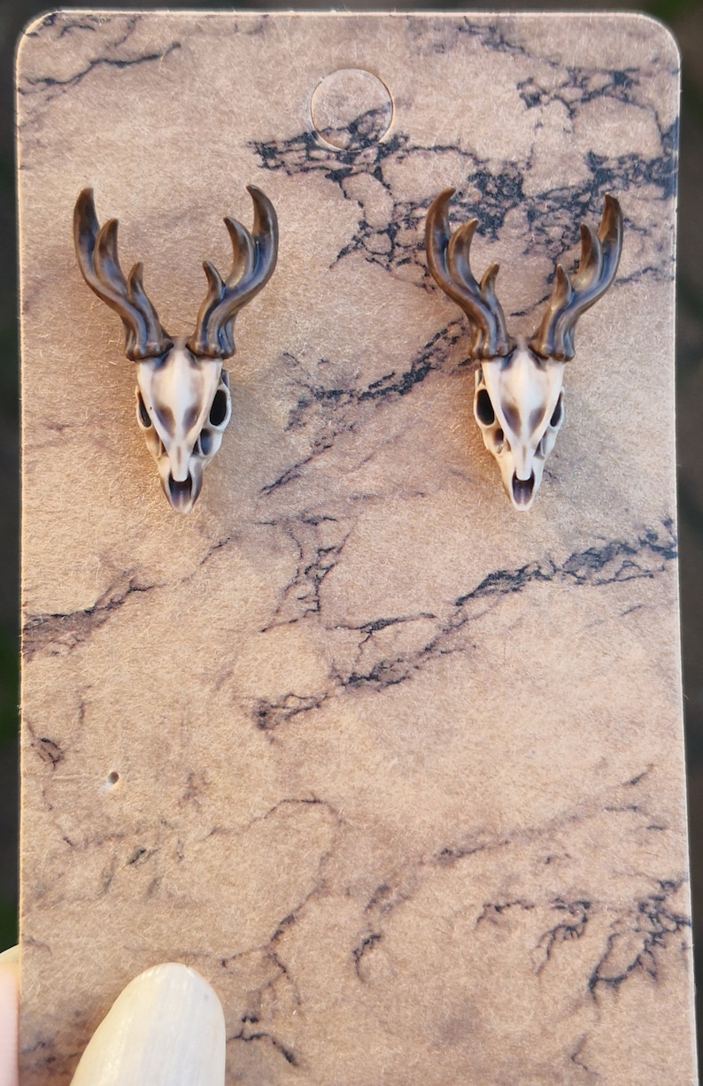 May include: A pair of brown and white deer skull earrings. The earrings are on a brown background.