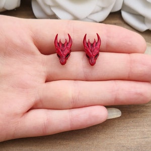 Pode incluir: Um par de brincos pequenos de cabe&ccedil;a de drag&atilde;o vermelhos. Cada brinco apresenta uma cabe&ccedil;a de drag&atilde;o detalhada com chifres e tra&ccedil;os faciais. Os brincos s&atilde;o projetados para serem usados como brincos de press&atilde;o.