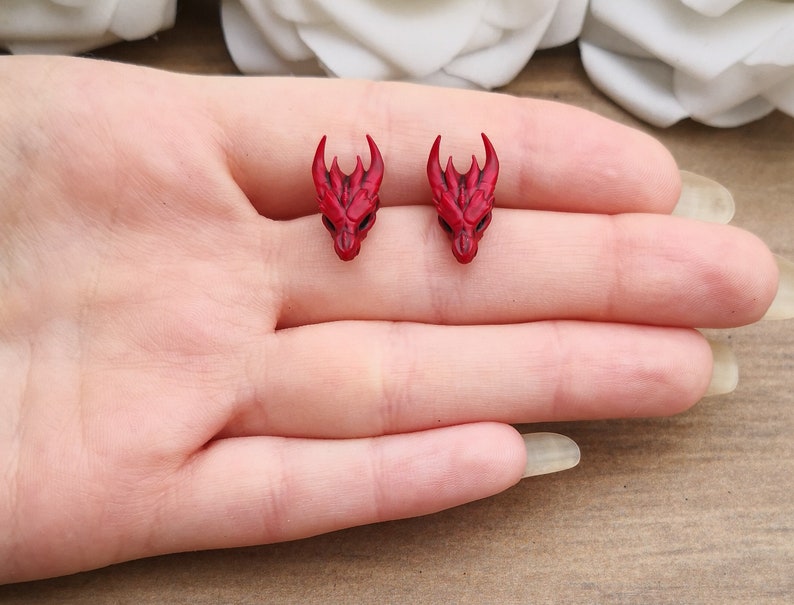 Pode incluir: Um par de brincos pequenos de cabe&ccedil;a de drag&atilde;o vermelhos. Cada brinco apresenta uma cabe&ccedil;a de drag&atilde;o detalhada com chifres e tra&ccedil;os faciais. Os brincos s&atilde;o projetados para serem usados como brincos de press&atilde;o.