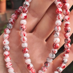 May include: A beaded bracelet featuring round white beads with a decorative pattern of red paint splatters and thin black lines. The beads are strung together in a continuous loop, creating a striking contrast between the white base and the dark markings.