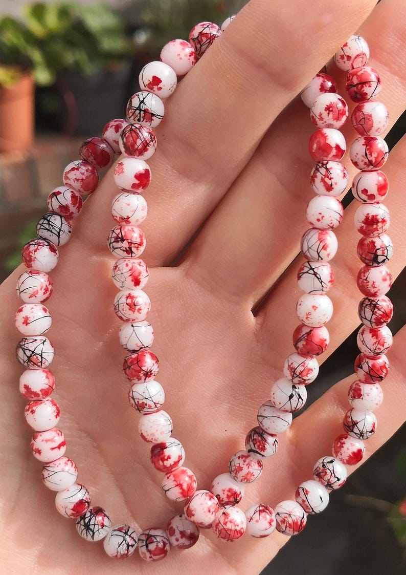 May include: A beaded bracelet featuring round white beads with a decorative pattern of red paint splatters and thin black lines. The beads are strung together in a continuous loop, creating a striking contrast between the white base and the dark markings.