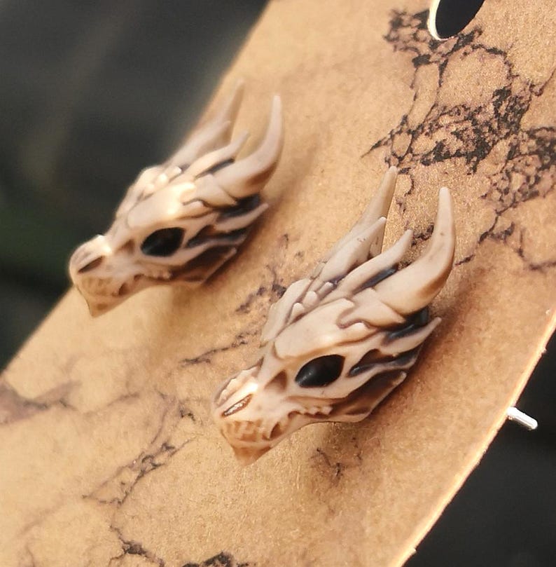 May include: A pair of dragon head stud earrings. The earrings are made of a white material with black details. The dragon heads are facing forward with their mouths open.
