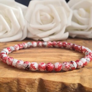 May include: A circular beaded bracelet featuring small, round beads with a white base color and irregular red splattered patterns, accented by thin black crackle lines throughout the surface of each individual bead in the strand.