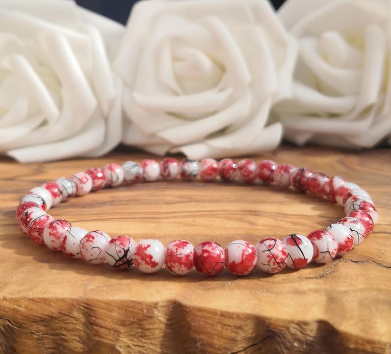 May include: A circular beaded bracelet featuring small, round beads with a white base color and irregular red splattered patterns, accented by thin black crackle lines throughout the surface of each individual bead in the strand.