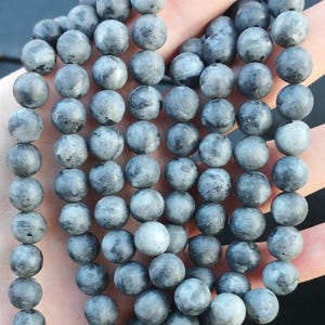 May include: A string of grey gemstone beads. The beads are round, with a matte finish and a mottled appearance. The beads are approximately 8mm in diameter. The strand is held in a hand.