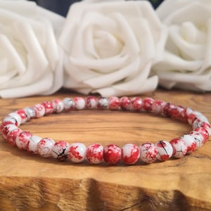 May include: A circular beaded bracelet featuring small, round white beads with a distinct red splatter pattern and thin black veining throughout. The beads are uniform in size and arranged in a continuous loop, creating a speckled appearance.