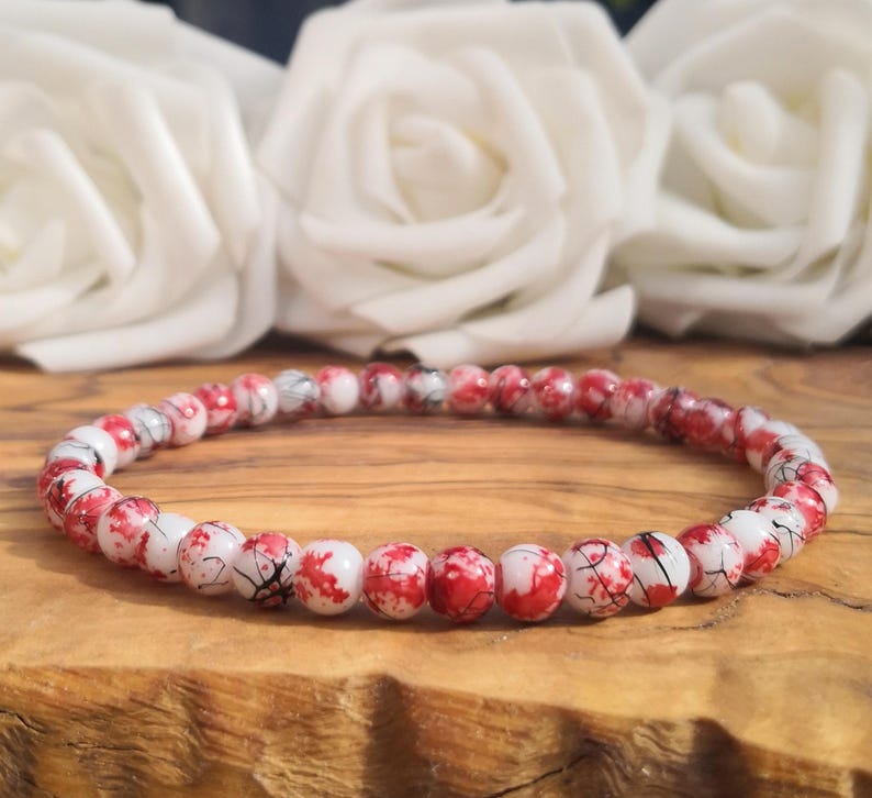 May include: A circular beaded bracelet featuring small, round white beads with a distinct red splatter pattern and thin black veining throughout. The beads are uniform in size and arranged in a continuous loop, creating a speckled appearance.