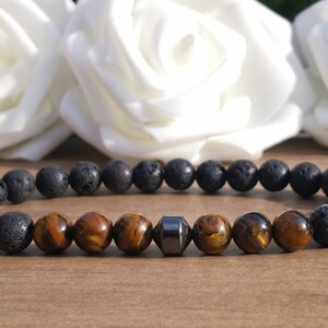 May include: A bracelet made of black lava rock beads, interspersed with tiger's eye beads and a single, dark grey, geometric bead. The bracelet is displayed against a soft background.