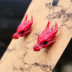 Pode incluir: Um par de brincos de press&atilde;o com cabe&ccedil;a de drag&atilde;o vermelha. Cada brinco apresenta uma cabe&ccedil;a de drag&atilde;o detalhada com chifres, olhos pretos e escamas texturizadas. Os brincos s&atilde;o exibidos em um cart&atilde;o marrom.