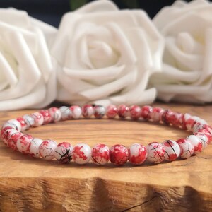 May include: A beaded bracelet featuring small, round, white beads with a vibrant red splatter pattern and fine black crackle veins throughout. The beads are strung together in a continuous circular loop, creating a uniform and detailed design.