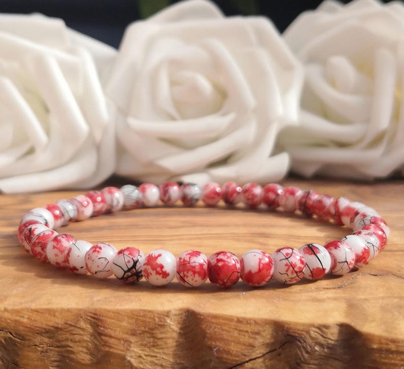 May include: A beaded bracelet featuring small, round, white beads with a vibrant red splatter pattern and fine black crackle veins throughout. The beads are strung together in a continuous circular loop, creating a uniform and detailed design.