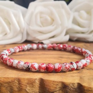 May include: A beaded bracelet featuring small, round beads with a white base and a mottled red and black marbled pattern. The beads are strung together in a continuous circular loop, creating a uniform design that displays a distinct color contrast.