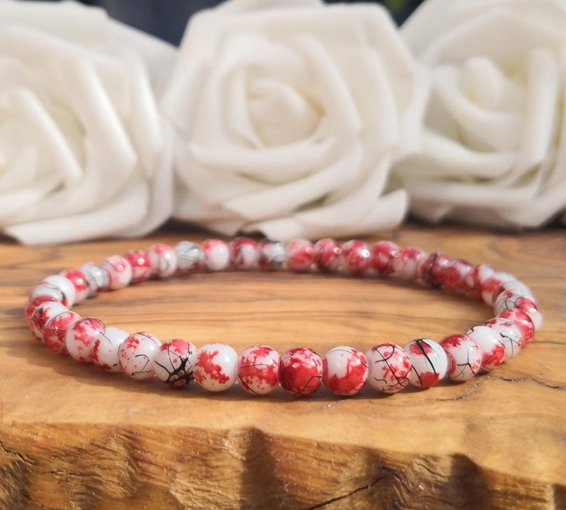 May include: A beaded bracelet featuring small, round beads with a white base and a mottled red and black marbled pattern. The beads are strung together in a continuous circular loop, creating a uniform design that displays a distinct color contrast.