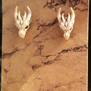 May include: A pair of white dragon head earrings with black detailing. The earrings are displayed on a brown card with a marbled pattern.
