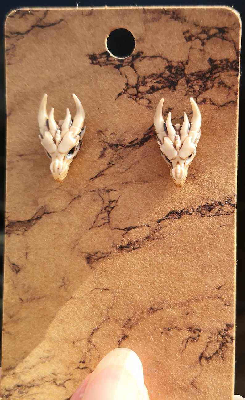 May include: A pair of white dragon head earrings with black detailing. The earrings are displayed on a brown card with a marbled pattern.