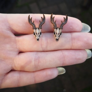 May include: A pair of deer skull earrings with brown antlers. The earrings are shown on a hand.