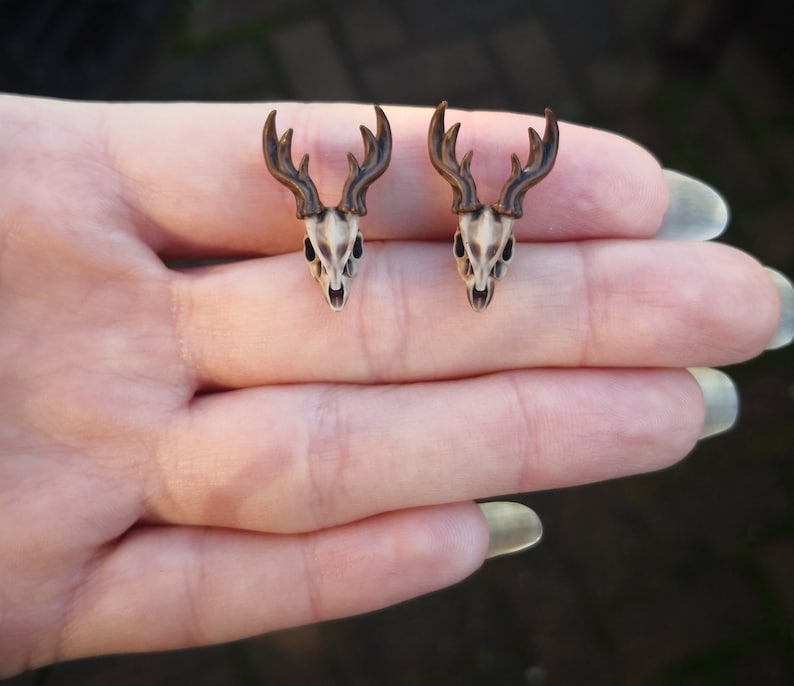 May include: A pair of deer skull earrings with brown antlers. The earrings are shown on a hand.