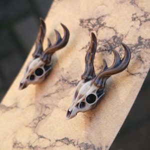 May include: A pair of deer skull earrings with antlers. The skulls are a light beige color with black eyes. The antlers are a dark brown color.