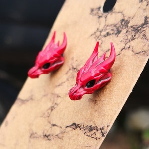 Pode incluir: Um par de brincos de press&atilde;o com cabe&ccedil;a de drag&atilde;o vermelha. Cada brinco apresenta um design detalhado de caveira de drag&atilde;o com chifres e &oacute;rbitas oculares pretas. Os brincos s&atilde;o exibidos em um cart&atilde;o castanho.