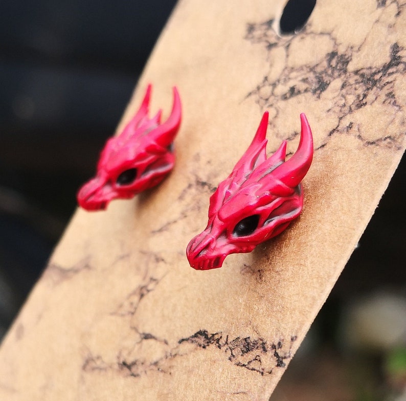 Pode incluir: Um par de brincos de press&atilde;o com cabe&ccedil;a de drag&atilde;o vermelha. Cada brinco apresenta um design detalhado de caveira de drag&atilde;o com chifres e &oacute;rbitas oculares pretas. Os brincos s&atilde;o exibidos em um cart&atilde;o castanho.