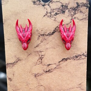 Pode incluir: Um par de brincos de press&atilde;o com cabe&ccedil;a de drag&atilde;o vermelha. Os brincos s&atilde;o feitos de um material vermelho brilhante com detalhes, incluindo chifres e estruturas faciais. Eles s&atilde;o exibidos em um cart&atilde;o marrom com um padr&atilde;o de m&aacute;rmore.
