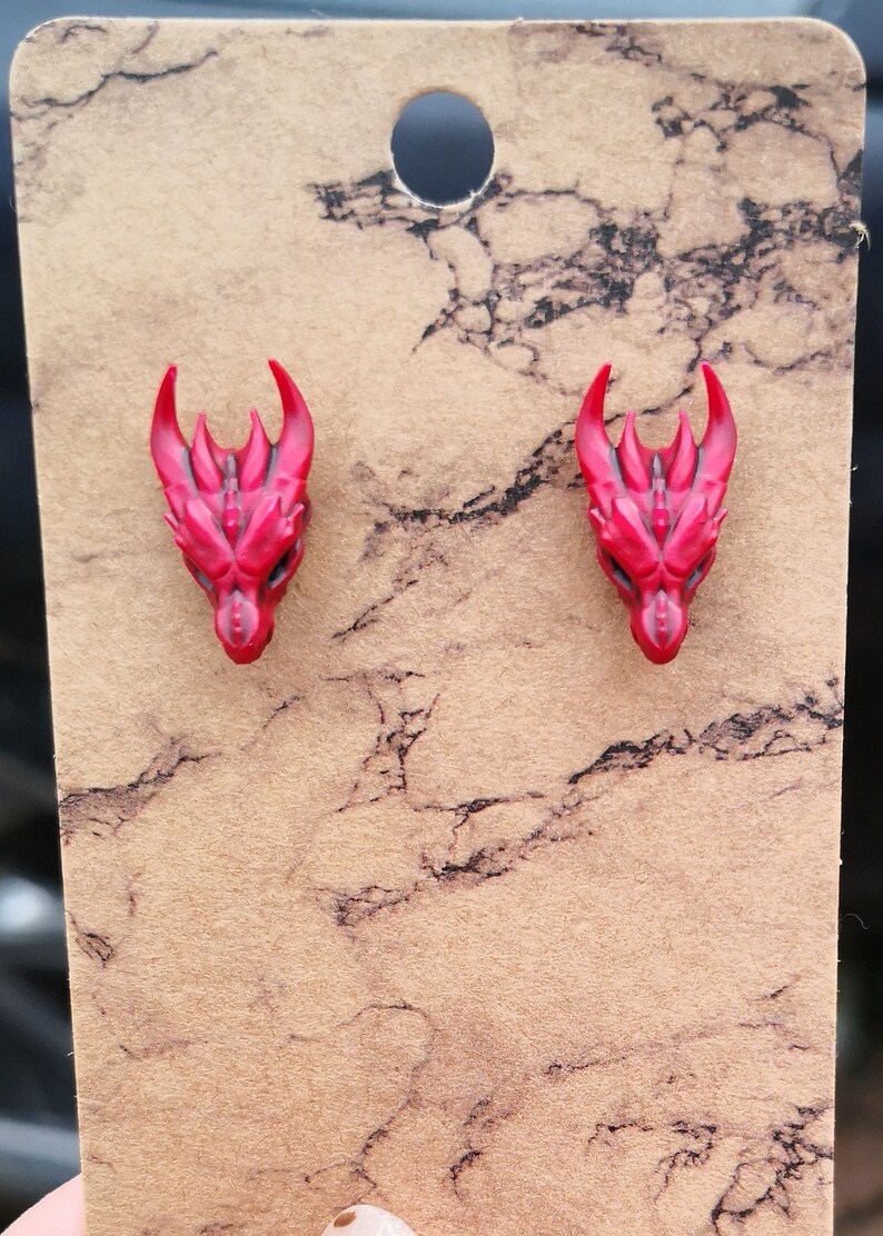 Pode incluir: Um par de brincos de press&atilde;o com cabe&ccedil;a de drag&atilde;o vermelha. Os brincos s&atilde;o feitos de um material vermelho brilhante com detalhes, incluindo chifres e estruturas faciais. Eles s&atilde;o exibidos em um cart&atilde;o marrom com um padr&atilde;o de m&aacute;rmore.