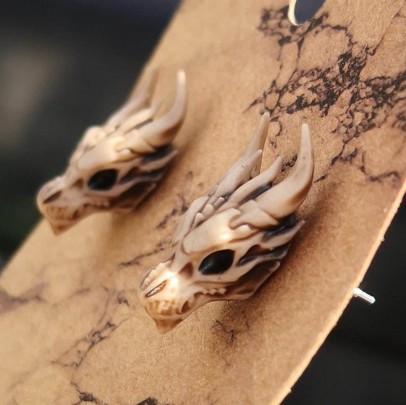 May include: A pair of dragon skull earrings made of white resin with black eyes. The earrings are displayed on a brown background.
