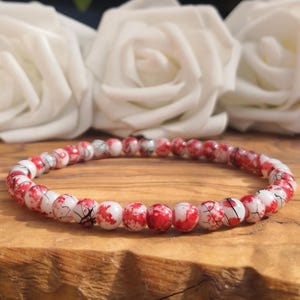 May include: A beaded bracelet featuring small, round, white beads with vibrant red splattered patterns and thin black crackle-like veins throughout. The beads are strung together in a continuous circular loop, creating a uniform and detailed design.