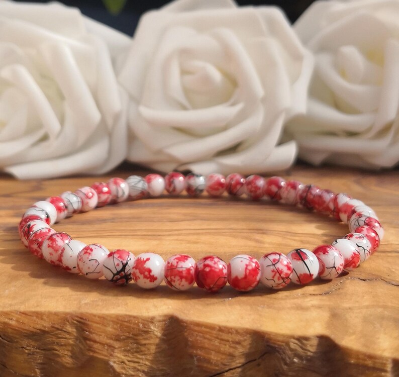 May include: A beaded bracelet featuring small, round, white beads with vibrant red splattered patterns and thin black crackle-like veins throughout. The beads are strung together in a continuous circular loop, creating a uniform and detailed design.