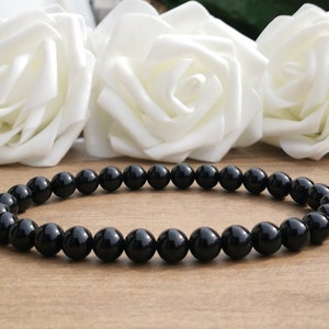 May include: A black beaded bracelet with polished, round beads. The bracelet is displayed on a light brown surface, with white roses in the background. The beads are evenly sized and have a glossy finish.