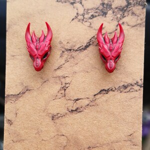 Pode incluir: Um par de brincos de press&atilde;o com cabe&ccedil;a de drag&atilde;o vermelha. Os brincos apresentam chifres e tra&ccedil;os faciais detalhados. S&atilde;o apresentados em um cart&atilde;o marrom com um padr&atilde;o de m&aacute;rmore. Um acess&oacute;rio com tema de fantasia.