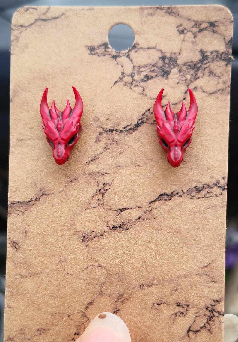 Pode incluir: Um par de brincos de press&atilde;o com cabe&ccedil;a de drag&atilde;o vermelha. Os brincos apresentam chifres e tra&ccedil;os faciais detalhados. S&atilde;o apresentados em um cart&atilde;o marrom com um padr&atilde;o de m&aacute;rmore. Um acess&oacute;rio com tema de fantasia.