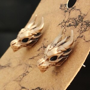 May include: A pair of dragon head stud earrings. The earrings are made of a light brown material and have black eyes.