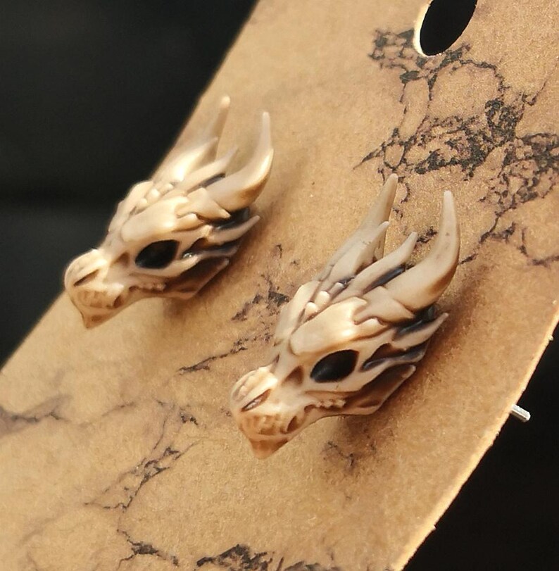 May include: A pair of dragon head stud earrings. The earrings are made of a light brown material and have black eyes.