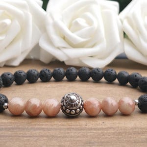 May include: A beaded bracelet featuring a series of porous black lava stone beads and six smooth, peach-coloured sunstone beads. A single, ornate, silver-toned metal bead with intricate embossed detailing is positioned in the centre of the sunstones.