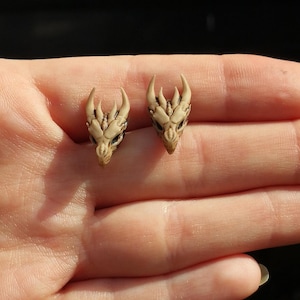 May include: A pair of dragon head earrings made of polymer clay. The earrings are a light beige colour and have detailed features.