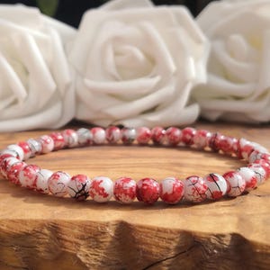 May include: A beaded bracelet featuring small, round, white beads with a vibrant red splatter pattern and fine black crackle detailing. The beads are strung together in a continuous circular loop, creating a uniform and detailed accessory.