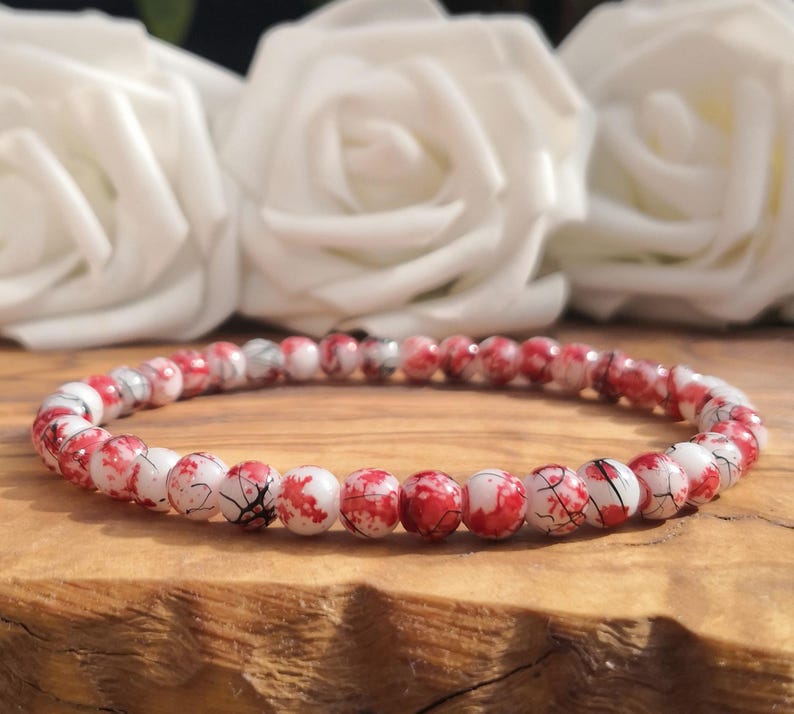 May include: A beaded bracelet featuring small, round, white beads with a vibrant red splatter pattern and fine black crackle detailing. The beads are strung together in a continuous circular loop, creating a uniform and detailed accessory.